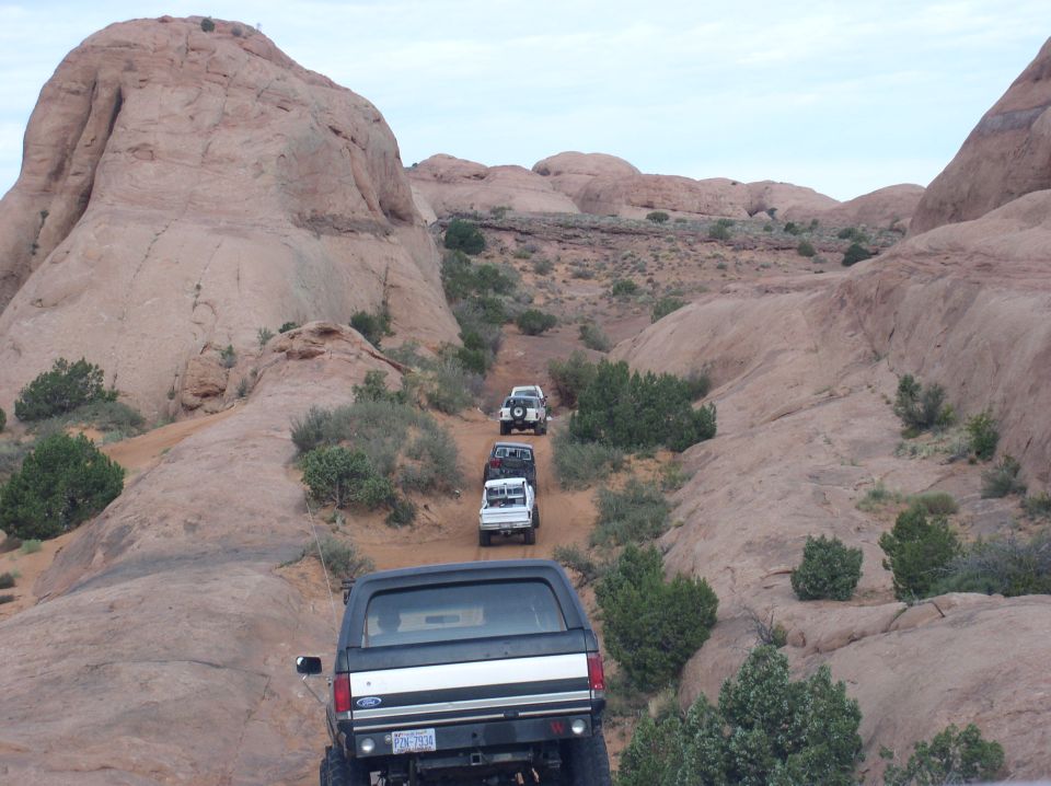 Four-wheel drive vehicles driving up a rocky trail in a desert landscape. The lead vehicle is a Ford Bronco
