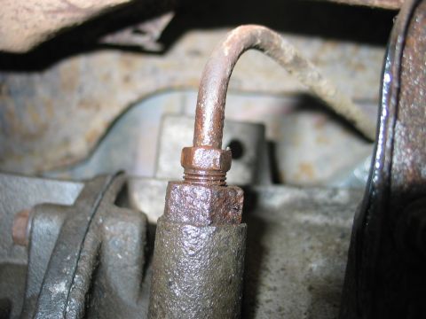 1995 Ford Bronco - Extreme close-up of a rusted hydraulic line where its hex fitting threads into a cast-iron component, with the tube curving up and out of f…