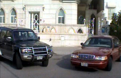 2000 Ford Crown Victoria - A maroon 2000 Ford Crown Victoria is parked in front of a large building, alongside a black SUV.