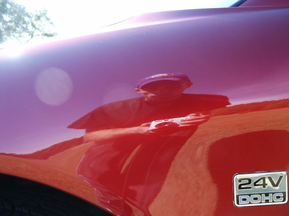 Red car fender with a reflection of a person holding a camera and a '24V DOHC' badge