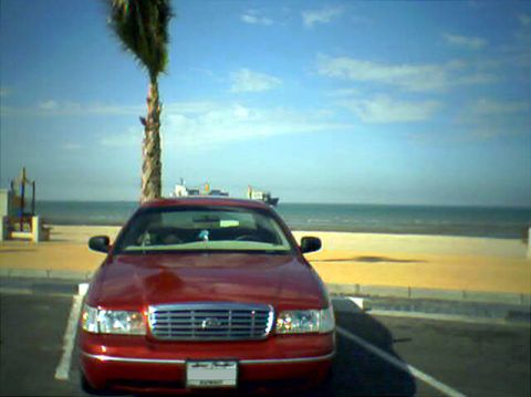 2000 Ford Crown Victoria - A red 2000 Ford Crown Victoria is parked facing the ocean on a sunny day, with a palm tree to its left.