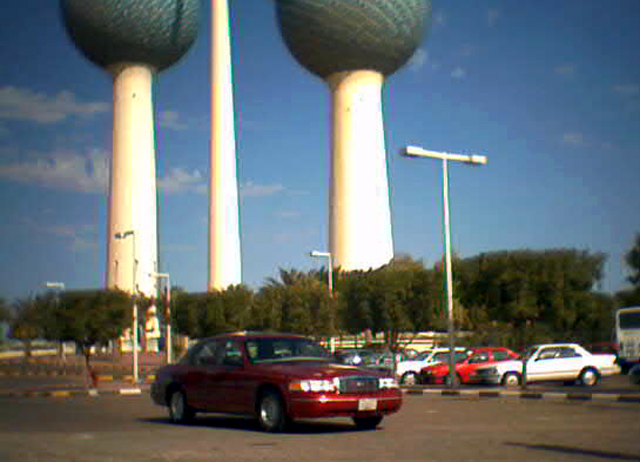 Red sedan parked in front of two large water towers. Other cars are in the background