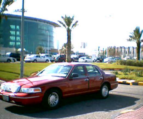 2000 Ford Crown Victoria - A red 2000 Ford Crown Victoria is parked in a lot with other vehicles and a modern building in the background.