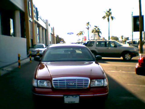 2000 Ford Crown Victoria - A red 2000 Ford Crown Victoria is parked on a street, with other vehicles and buildings visible in the background.