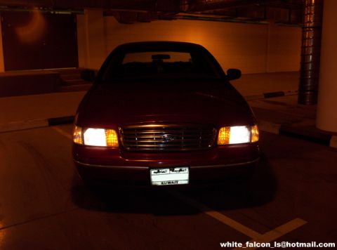 2000 Ford Crown Victoria - A red 2000 Ford Crown Victoria is parked indoors, facing the camera with its headlights illuminated.