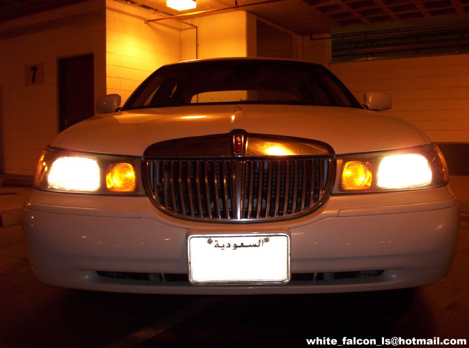 White Lincoln Town Car front view, grille with chrome accents, illuminated headlights and amber turn signals, Saudi Arabian license plate