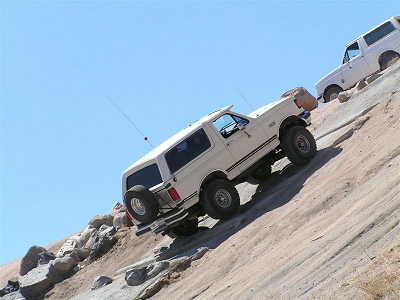 1988 Ford Bronco - Two white Ford Broncos ascend a challenging off-road trail under a clear blue sky.