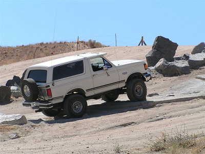 1988 Ford Bronco - A white 1988 Ford Bronco navigates a rocky off-road course under a clear blue sky.