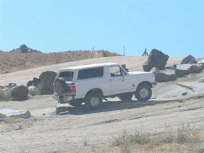 1988 Ford Bronco - A white Ford Bronco navigates a rocky, off-road terrain under a clear blue sky.