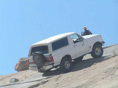 1988 Ford Bronco - A white Ford Bronco is driven uphill on a rocky surface under a clear blue sky.