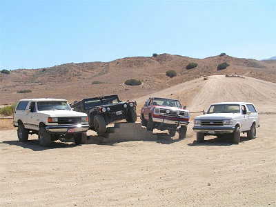 1988 Ford Bronco - Four off-road vehicles are parked in a desert landscape under a clear blue sky.