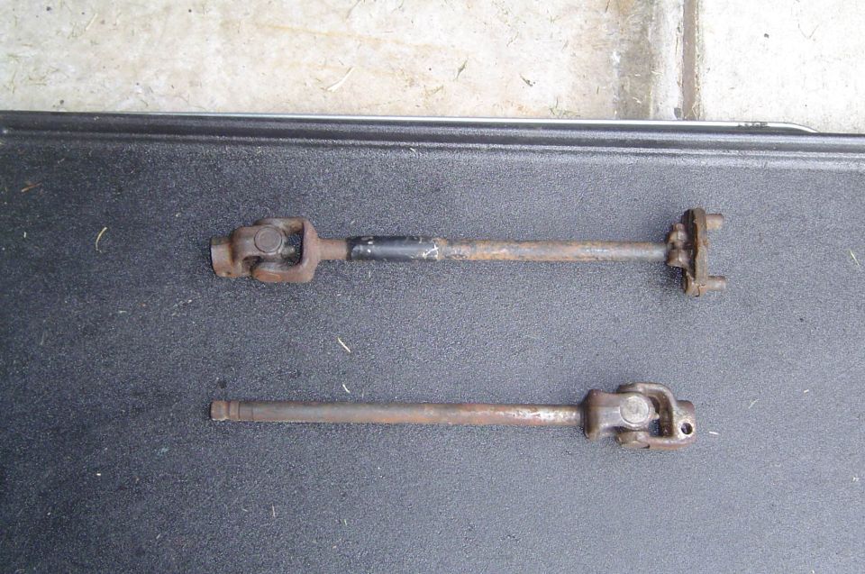 Two rusty steering shafts with U-joints laid side by side on a black bedliner for comparison
