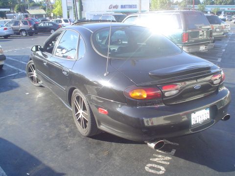 1993 Ford Taurus - This black Ford Taurus sedan is parked in a lot, showing its rear end with a spoiler and dual exhaust pipes.