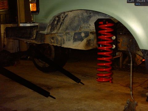 1995 Ford Bronco - Front suspension work on a light green Ford Bronco XL in a garage: the wheel and axle are dropped out, a tall red lift coil spring hangs fr…