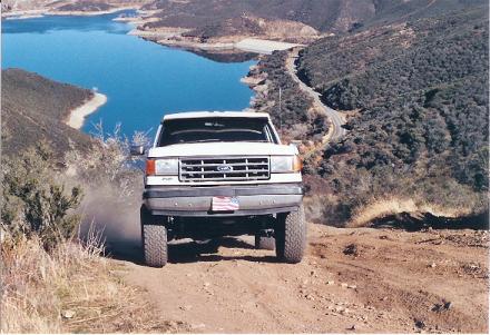 1988 Ford Bronco - This white Ford Bronco is captured in a front-facing view as it navigates a dusty off-road trail, with a large body of water visible in the…