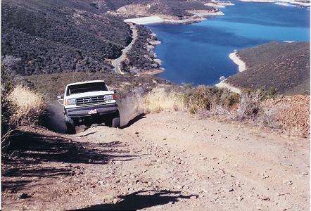 1988 Ford Bronco - A white Ford F-Series truck navigates a steep, dusty off-road trail with a scenic lake in the background.