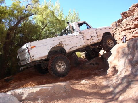 1993 Ford Bronco - A white pickup truck with significant body damage is shown climbing over large rocks on a sunny day.