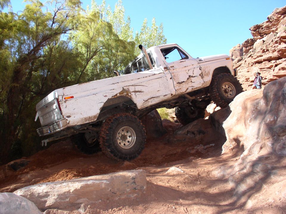 Muddy white Ford pickup with big mud tires and exhaust stack climbing a red rock ledge