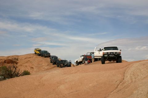 1993 Ford Bronco - A line of off-road vehicles, including a white 1993 Ford Bronco, navigates a rocky desert landscape under a cloudy sky.
