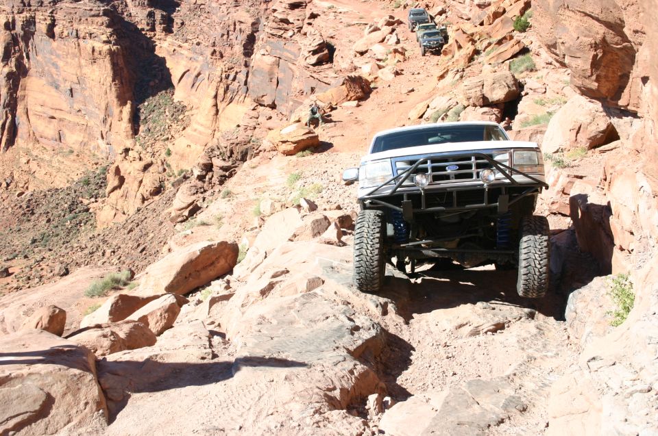 White lifted pickup with rear roll cage crawling over red sandstone ledges in a desert canyon