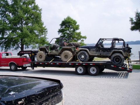 1992 Ford Bronco - A red pickup truck tows a trailer carrying two muddy off-road vehicles near a body of water.