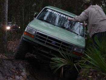 1994 Ford Bronco - A person cleans the windshield of a green Ford Bronco as it navigates a muddy, wooded trail. Another vehicle is visible in the background.