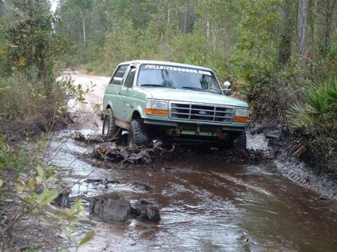 1994 Ford Bronco - A light green Ford Bronco navigates a muddy path through a wooded area.