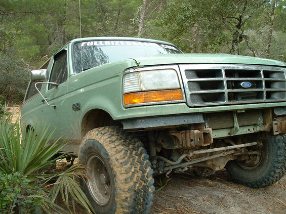 Olive-green Ford 4x4 with no front bumper flexing over roots on a sandy pine trail