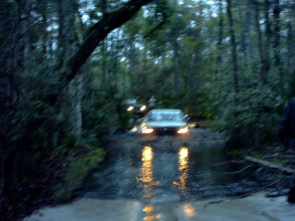 Ford 4x4 fording a flooded wooded trail at dusk with headlights reflecting on water