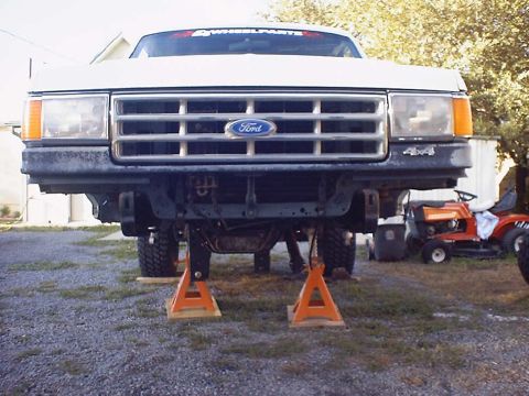 1988 Ford Bronco - Front suspension of a 1988 Ford Bronco is visible as the vehicle is supported by jack stands, with a riding mower in the background.