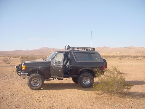 1988 Ford Bronco - A dark-colored Ford Bronco sits in a desert environment with its driver's door ajar and a roof rack installed.
