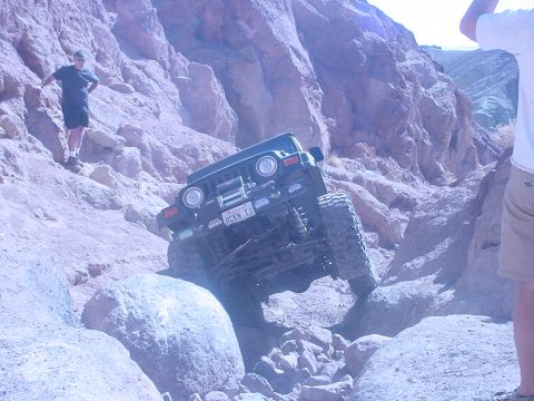 1988 Ford Bronco - A Jeep Wrangler navigates a steep, rocky canyon trail with a person standing on a ledge above.