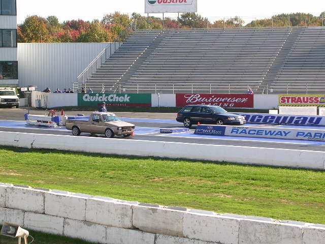 Tan pickup truck and dark station wagon on a drag strip with grandstands in the background
