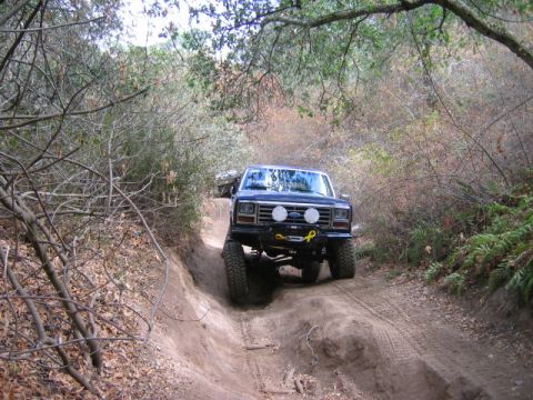 1985 Ford Bronco - A dark blue Ford Bronco navigates a steep dirt trail surrounded by dense foliage.