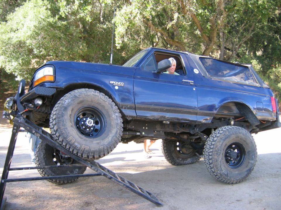 Lifted dark blue Ford Bronco on big mud tires climbing a ramp outdoors.
