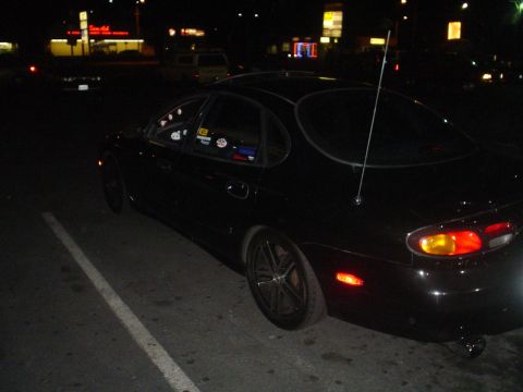 1993 Ford Taurus - A black sedan is parked in a lot at night, showing its rear quarter panel and a prominent antenna.