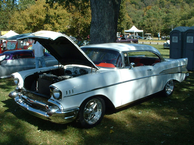 White 1957 Chevrolet Bel Air coupe with hood open, parked on grass at an outdoor car show. Another car is visible to the left