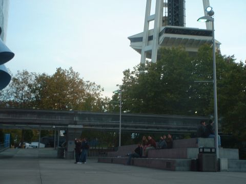 1999 Ford Crown Victoria - A view of the Seattle Space Needle with a monorail track overhead and people relaxing on tiered seating.