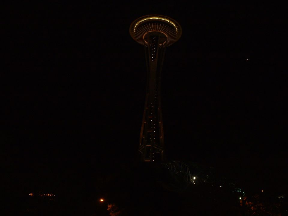Space Needle at night with lights on, dark sky, and a faint light in the distance