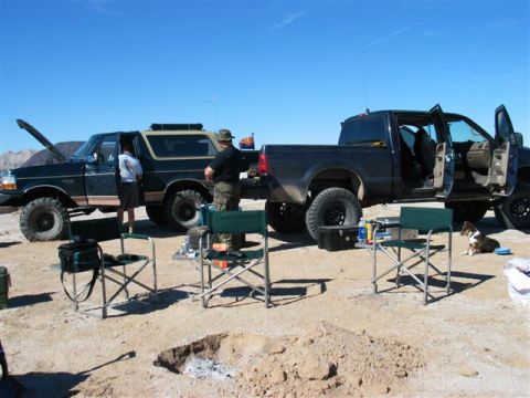 1994 Ford Bronco - Two off-road vehicles, a Bronco and a pickup truck, are parked in a desert setting with camping gear.