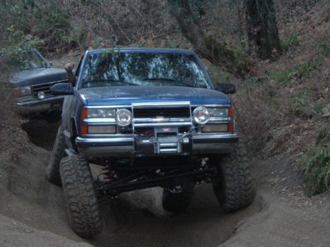 1989 Ford Bronco - A blue Chevrolet pickup truck with a winch is shown off-roading on a dirt path, with another vehicle following closely.