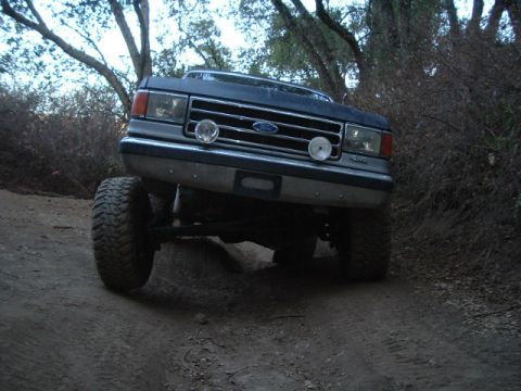 1989 Ford Bronco - This lifted Ford truck is navigating a rough, wooded trail with its front wheels angled.