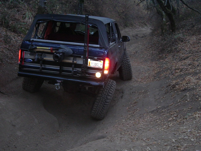 Dark blue Ford Bronco with soft top and tailgate jack rack climbing a dirt trail at dusk