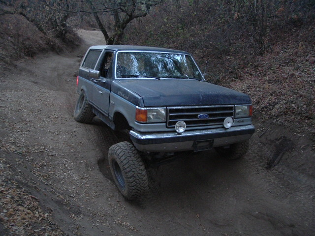 Lifted blue and silver Ford Bronco on a rutted dirt trail, front three-quarter view