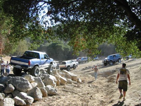 1985 Ford Bronco - Multiple off-road vehicles navigate a challenging dirt path with large rocks and trees.