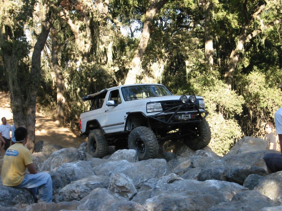 White lifted Ford Bronco rock crawling over boulders on a wooded off-road trail