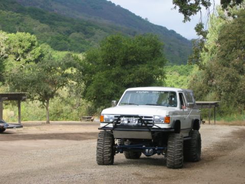 1985 Ford Bronco - Modified pickup truck with oversized tires and custom bumper sits in a rural, wooded area.