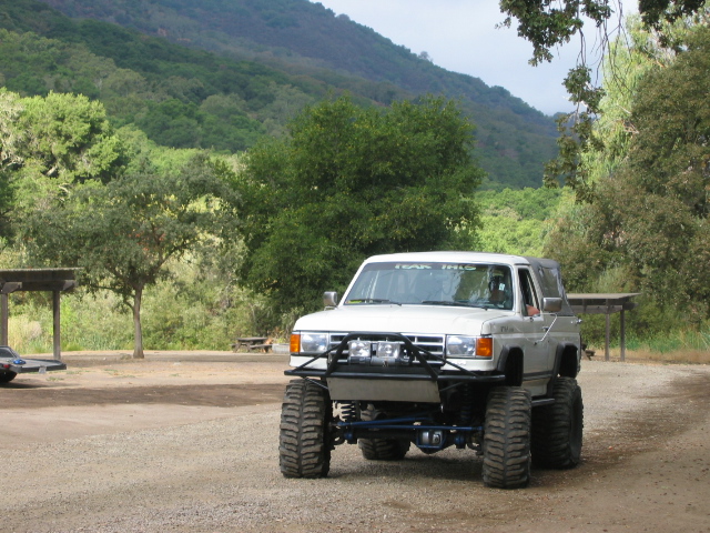 White lifted Ford Bronco on big off-road tires parked on gravel with green hills behind.