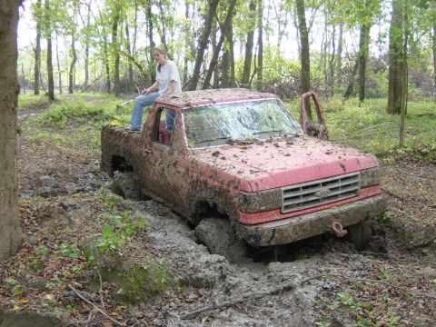1983 Ford Bronco - A woman sits atop a mud-covered pickup truck that is deeply stuck in a wooded area.