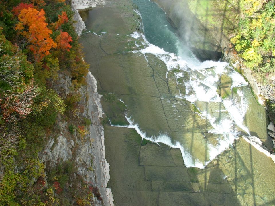 Waterfall cascading over layered rocks, with a cliff face covered in trees and autumn leaves on the left